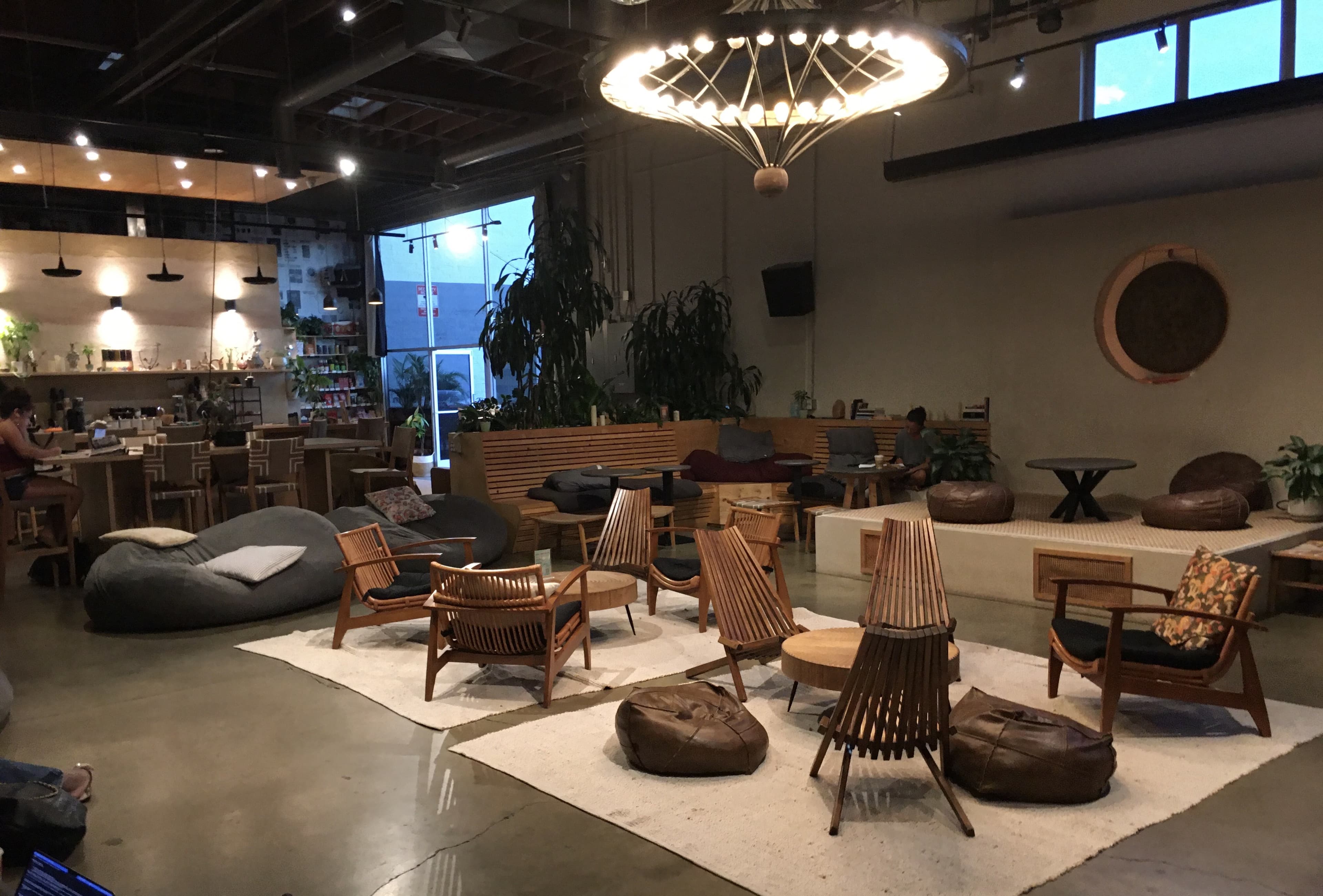 The main room at MUD\WTR: gather — armchairs drawn into a circle on a rug, under a wide pendant light, with plants and a café counter behind.
